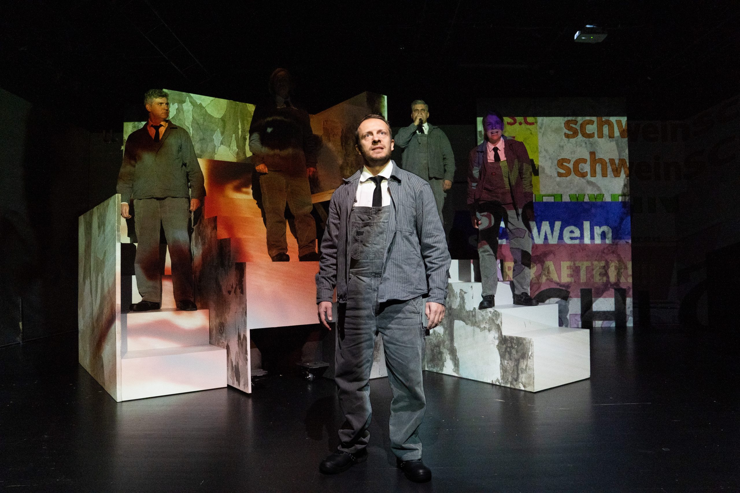 Fünf Personen in einem Bühnenbild aus vielen Treppen, darauf eine Projektion aus Wörtern