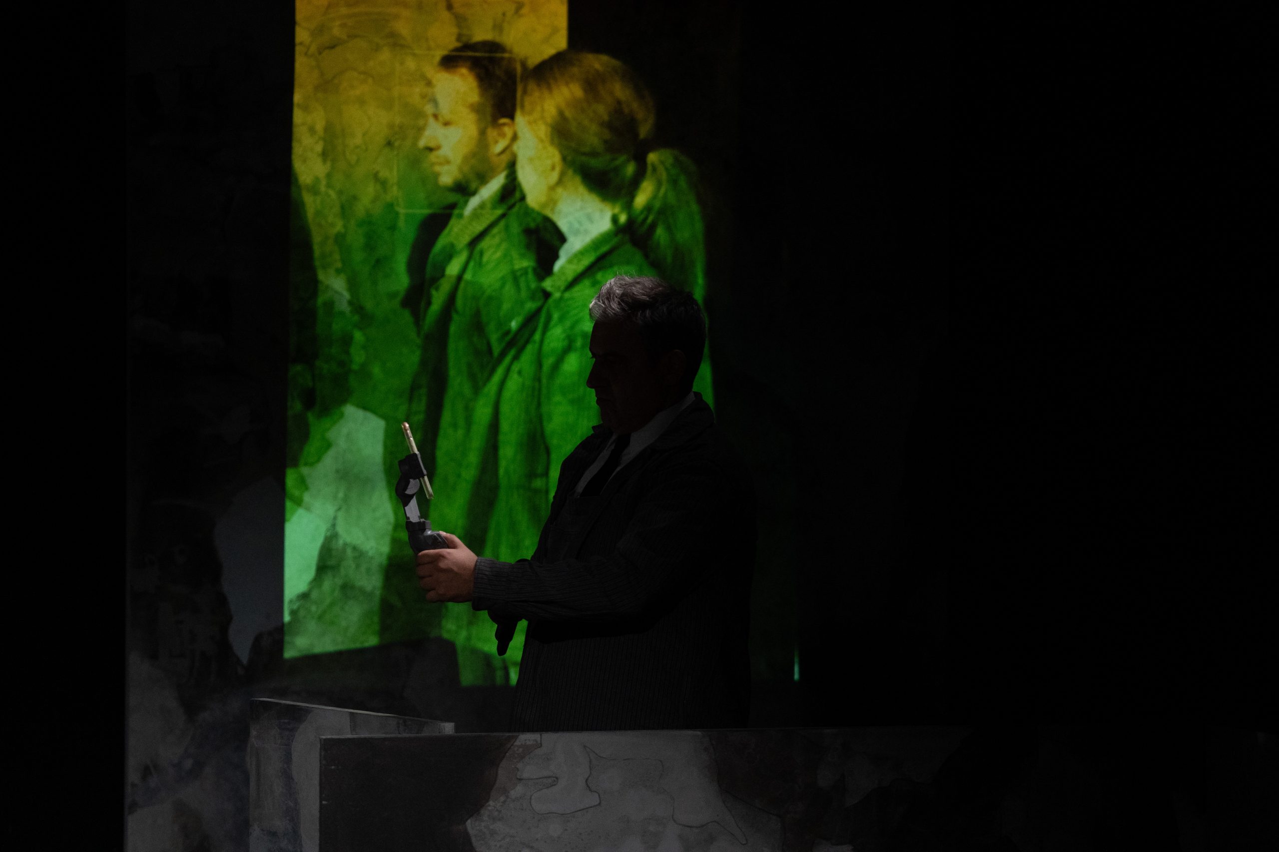 Eine Projektion vin zwei Menschen im Hintergrund, davor die Silouhette einer Person mit Handykamera