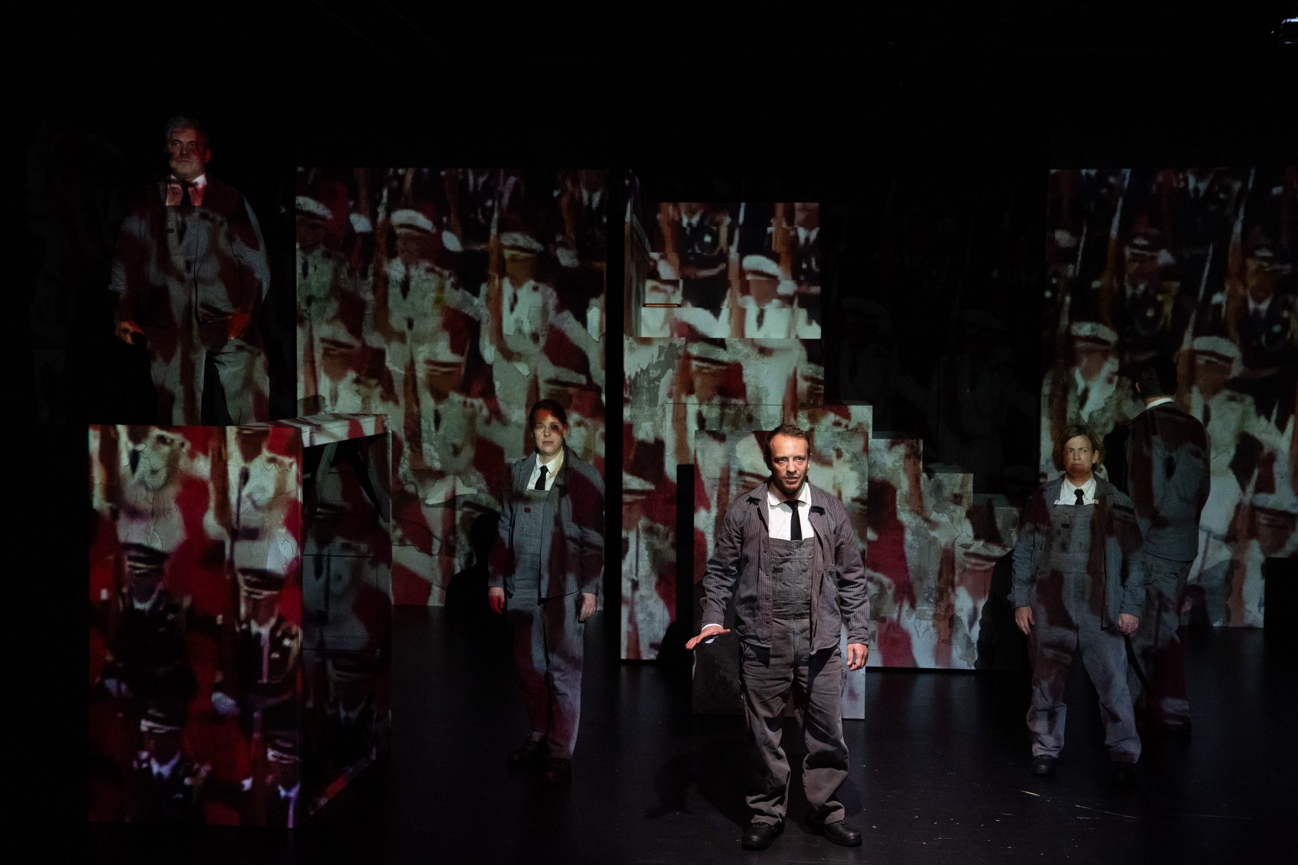 Personen auf einer Bühne, die von einer Projektion angestrahlt werden, die eine Armee zeigt