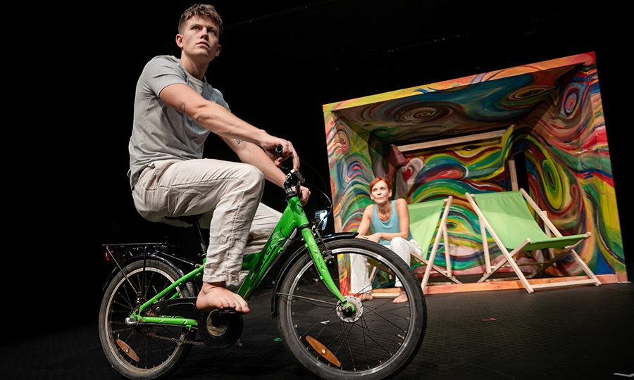 Ein junger Mann fährt auf einem Fahrrad, dahinter sitzt in einem bunt bemalten Verschlag eine Frau auf einem Campingstuhl