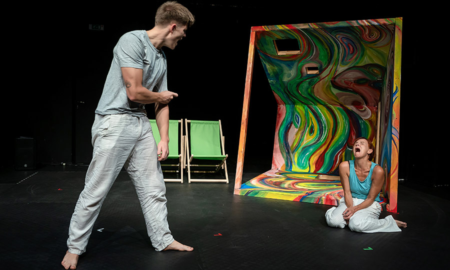 Eine Frau kniet vor einem bunt bemalten Verschlag und schreit, vor ihr steht ein junger Mann und schreit sie an