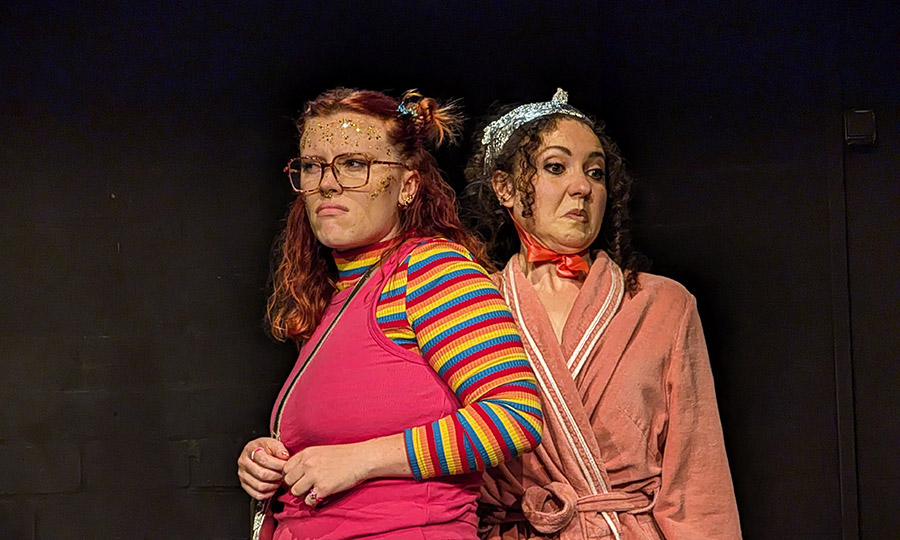 Eine Frau mit Glitzer im Gesicht guckt skeptisch, neben ihr steht eine Frau in Bademantel und mit Alufolie auf dem Kopf, dei angeekelt guckt.