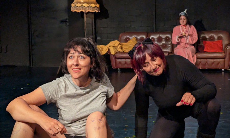 Eine Frau mit angemalten Beinen sitzt vor einer Couch und einem Lampenschirm. Ihren Arm legt sie auf die Schultern einer anderen Person mit Katzenohren.