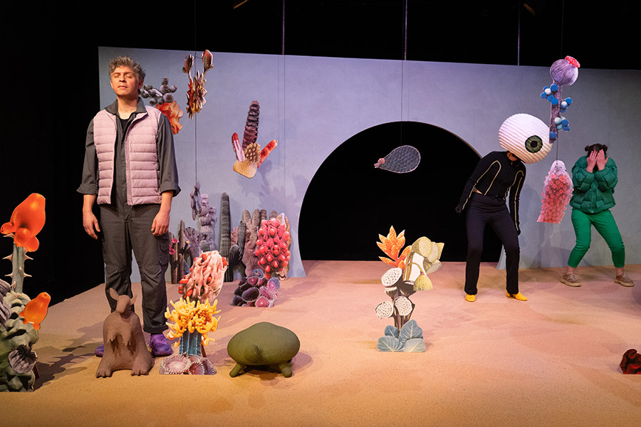 Zwischen Wüstenpflanzen steht eine Person mit entspanntem Gesichtsausdruck und geschlossenen Augen. Im Hintergrund ist eine Art Monster mit Riesemkopf und einem Auge zu sehen, daneben eine weitere Person, die sich die Augen zuhält.