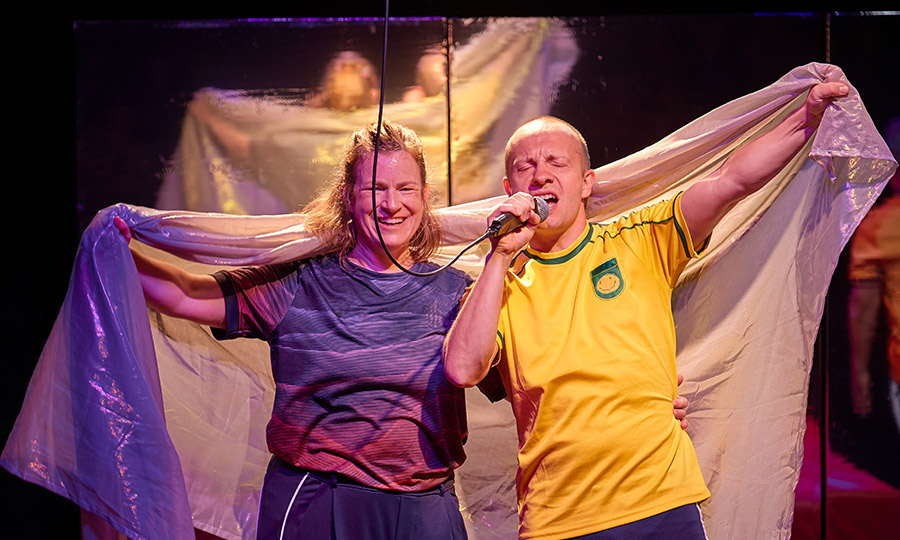 Zwei Personen in Sportkleidung halten ein großes weißes glitzerndes Tuch hoch. Die Person rechts singt in ein Mikro, die andere Person lächelt.