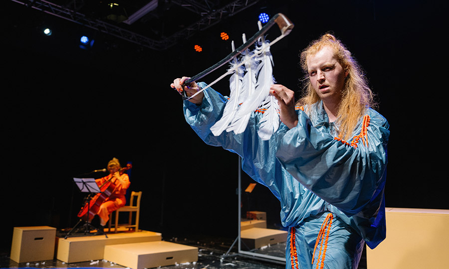 Eine Person in blauem Trainingsanzug hält einen Geigenbogen in die Höhe, an dem Federn hängen