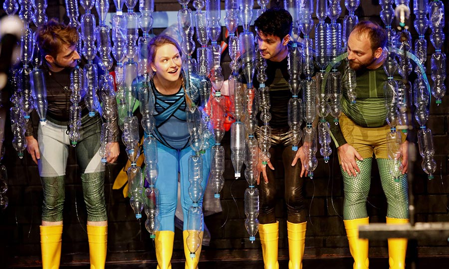 Vier Personen in gelben Gummistiefeln gucken durch eien Vorhang aus durchsichtigen Plastikflaschen