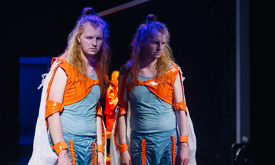 Eine Person mit orange-blauem Kostüm spiegelt sich in einer Scheibe