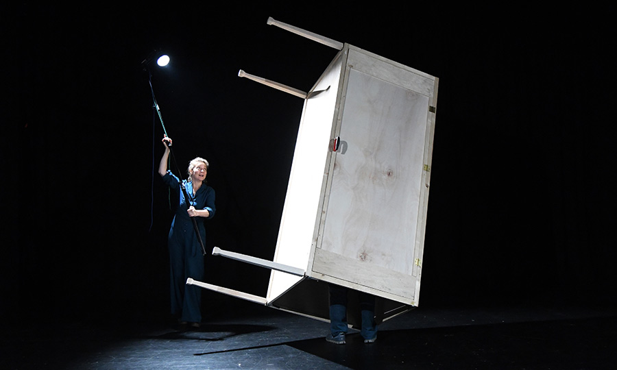Eine rechteckige große Holzbox steht hochkant nd schief auf einer Bühne, während eine Frau im Blaumann eine Lampe auf sie hält