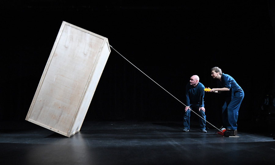 Eine große Holzbox steht schief auf einer Bühne und wird nur von einem dünnen Faden gehalten