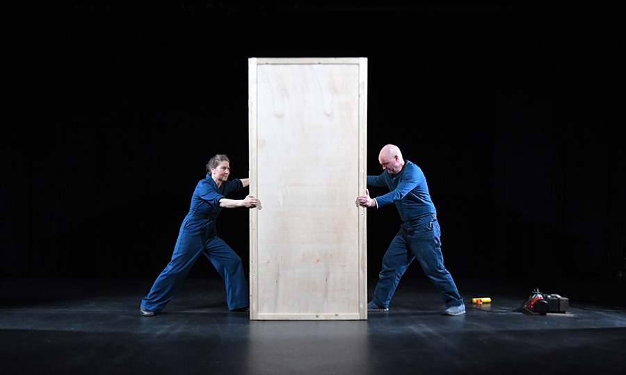 Zwei Menschen im Blaumann schieben eine große Holzbox über eine Bühne