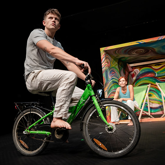 Ein junger Mann fährt auf einem Fahrrad, dahinter sitzt in einem bunt bemalten Verschlag eine Frau auf einem Campingstuhl