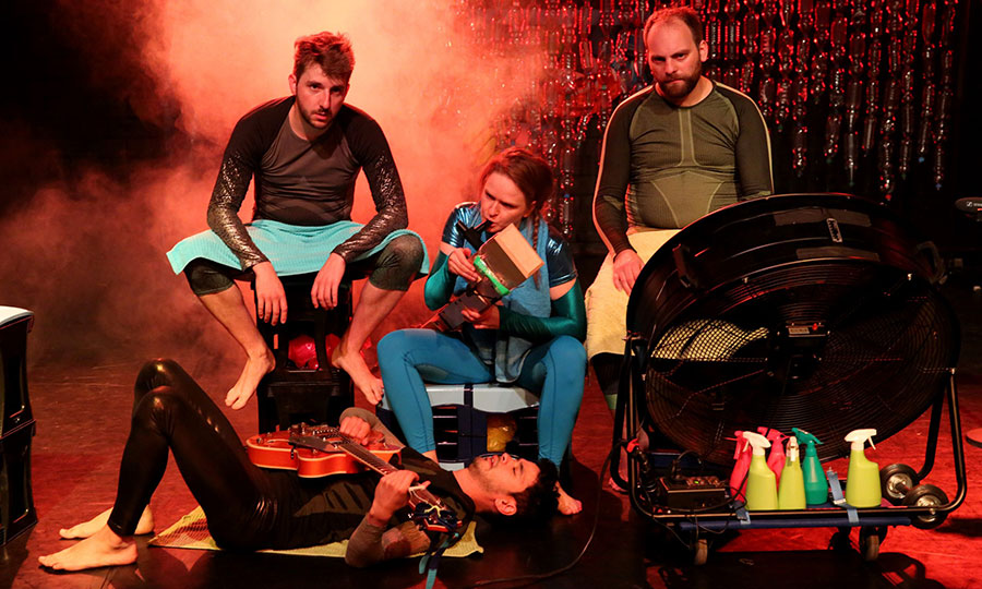 Vier Menschen sitzen bzw. liegen auf einer in rotes licht getauchten Bühne, hinter ihnen steigt Dampf auf