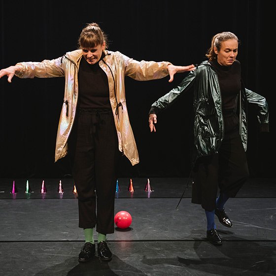 Zwei Menschen auf einer Bühne in zu großen Jacken, die die Arme ausbreiten