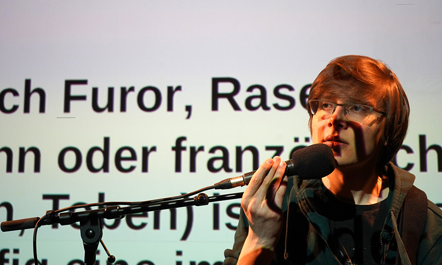 Eine Person mit Mikro sitzt vor einer Leinwand, auf die ein Text projiziert ist