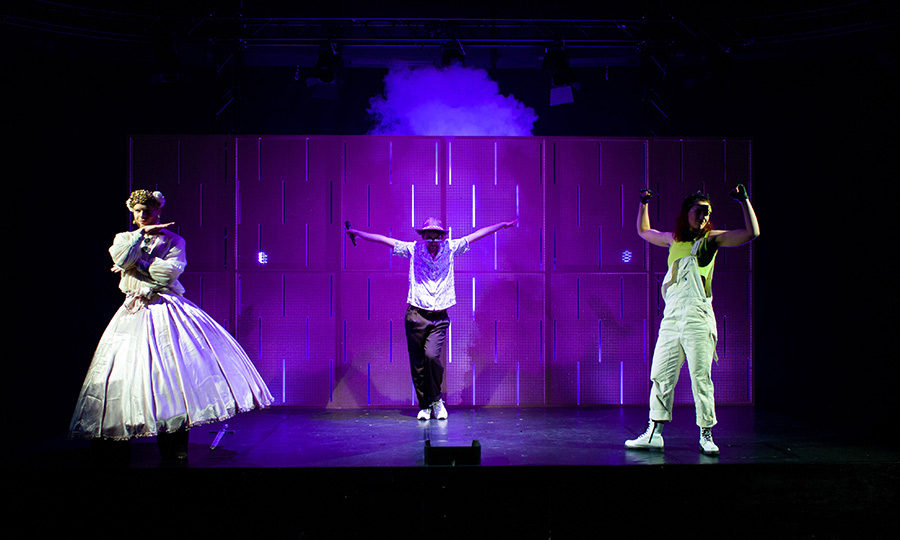 Drei Menschen auf einer Bühne, eine im weißen langen Kleid, in der MItte eine Person mit weißer Bluse und erhobenen Armen, rechts eine Person in Latzhose und ebenfalls mit erhobenen Armen