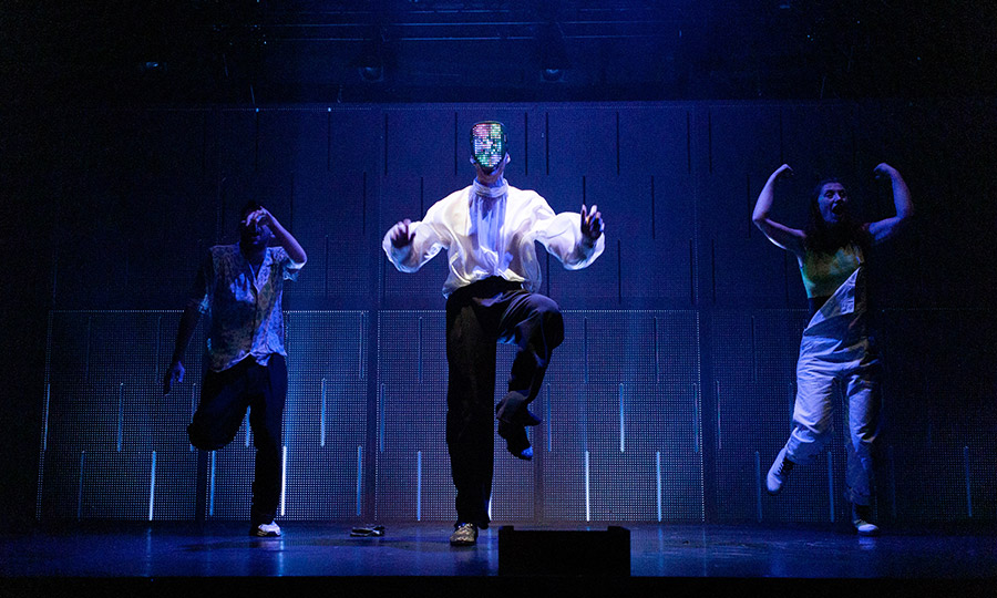 Drei Menschen hüpfen auf einer in blaues Licht getauchten Bühne, der mittlere trägt eine Maske vor dem Gesicht