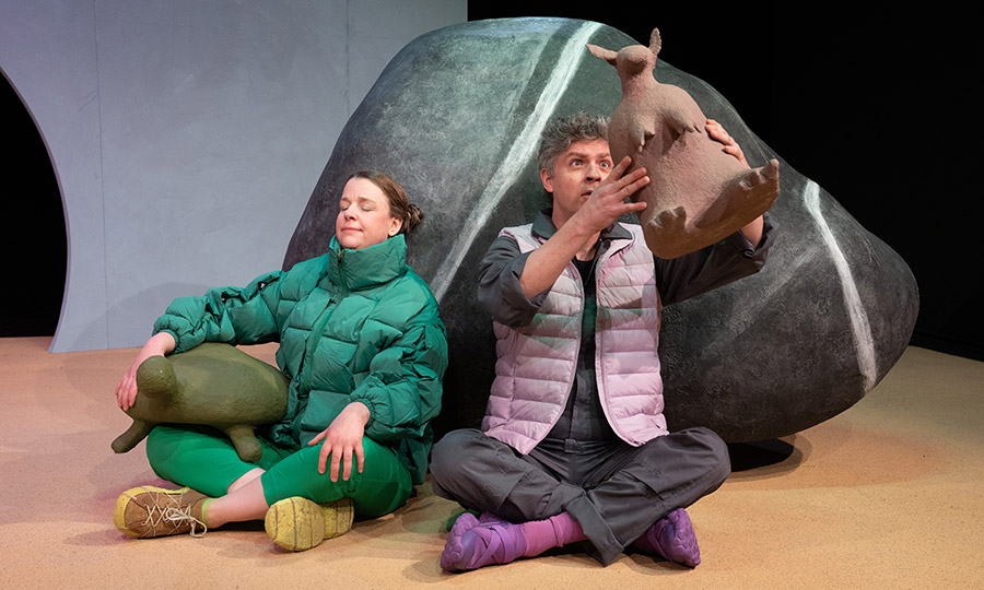 Zwei Personen sitzen vor einem großen Stein. Die rechte Person hält ein Gürteltier aus Pappmaché hoch.