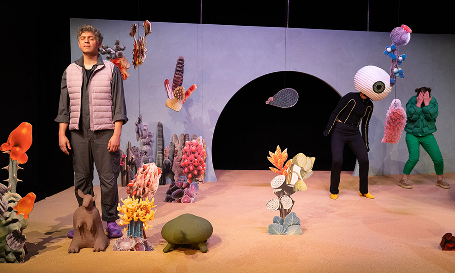 Zwischen Wüstenpflanzen steht eine Person mit entspanntem Gesichtsausdruck und geschlossenen Augen. Im Hintergrund ist eine Art Monster mit Riesemkopf und einem Auge zu sehen, daneben eine weitere Person, die sich die Augen zuhält.