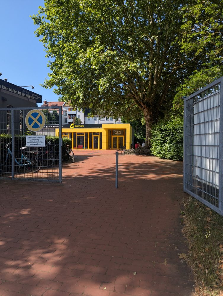 Der Eingang zum Hof des überzwerg. Im Vordergrund ein offenes Tor, dahinter ein rot gepflasteter Hof und im Hintergrund ein gelbes Gebäude.