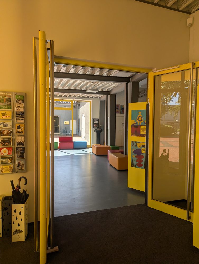 Der Eingang zum Foyer des überzwerg – Theater am Kästnerplatz: zwei offene Glastüren, dahinter ein Foyer mit Sitzgelegenheiten.