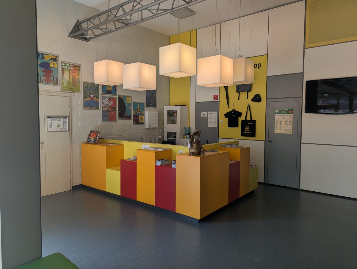Der Kassenbereich des überzwerg – Theater am Kästnerplatz: Eine Theke in orange, gelb und rot, darüber rechteckige Hängelampen, im Hintergrund sind Plakate von Stücken an den Wänden.